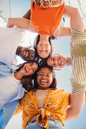 Vertical low view angle of a group of multiracial teenagers smiling and looking at camera together. Portrait of five young students smiling and laughing. Cheerful people having fun and embracing Vertical low view angle of a group of multiracial teenagers smiling and looking at camera together. Portrait of five young students smiling and laughing. Cheerful people having fun and embracing. High quality photo