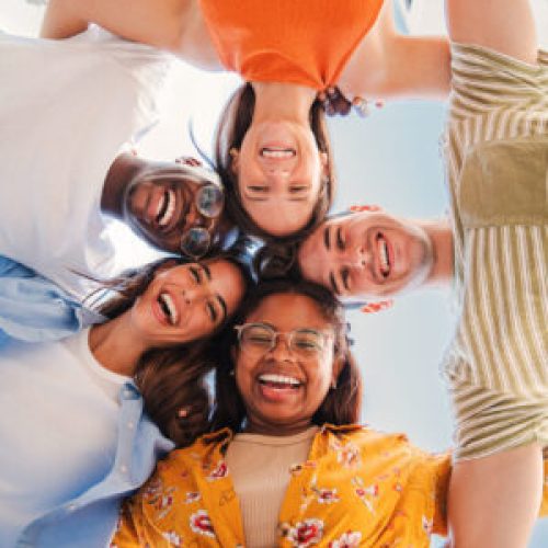 Vertical low view angle of a group of multiracial teenagers smiling and looking at camera together. Portrait of five young students smiling and laughing. Cheerful people having fun and embracing. High quality photo